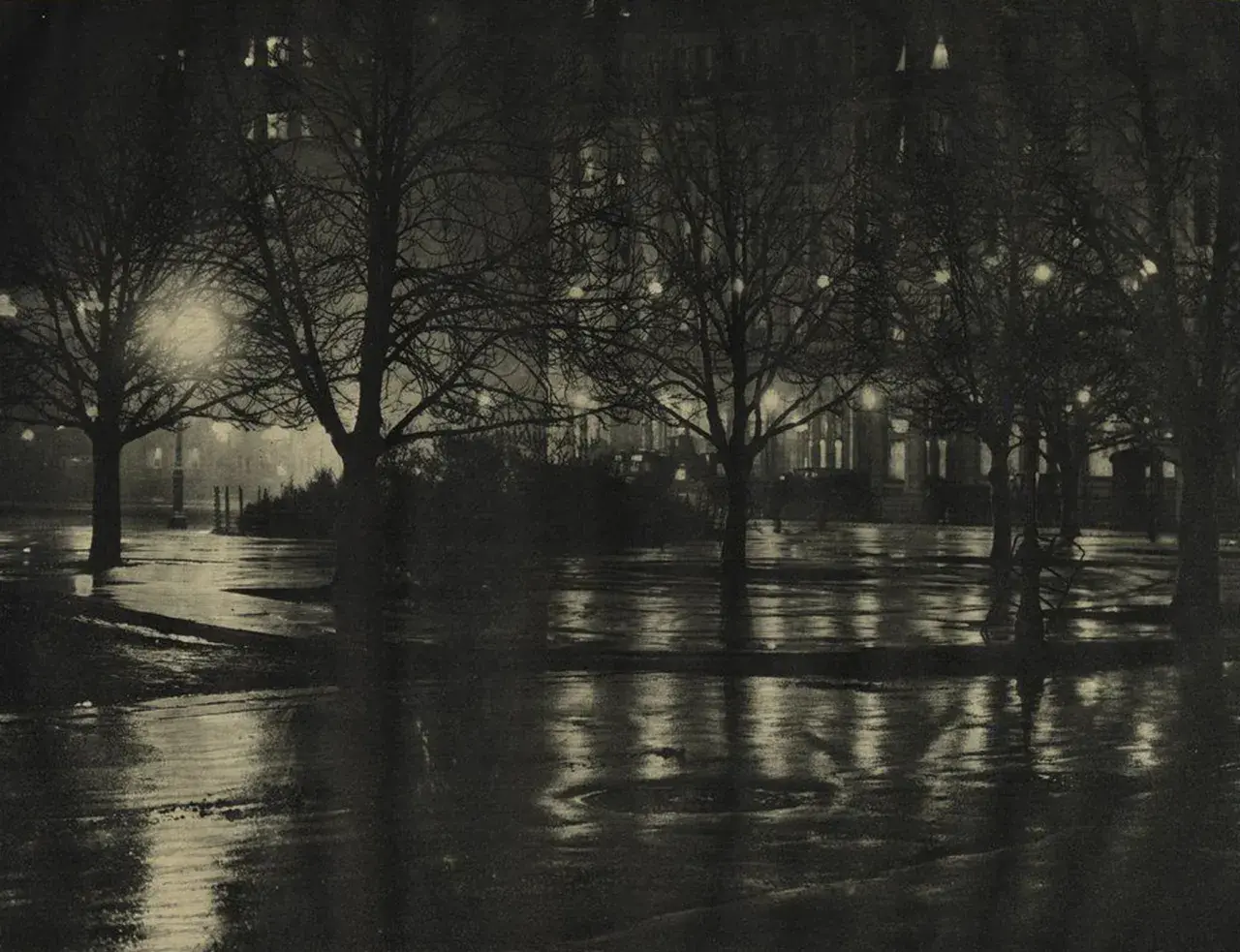 Nocny park po deszczu, drzewa bez liści, mokre ścieżki odbijające światła budynków. Alfred Stieglitz uchwycił atmosferę miasta.