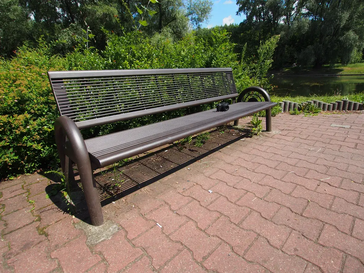 Szeroki kadr ukazuje pustą, metalową ławkę w parku, obok której leży aparat fotograficzny. W tle zielone drzewa i staw.