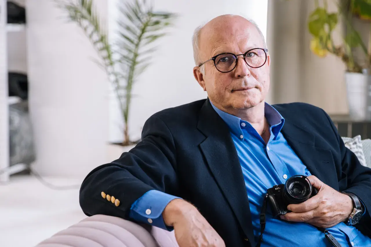 Tomasz Tomaszewski, w okularach i granatowej marynarce, trzyma aparat fotograficzny.