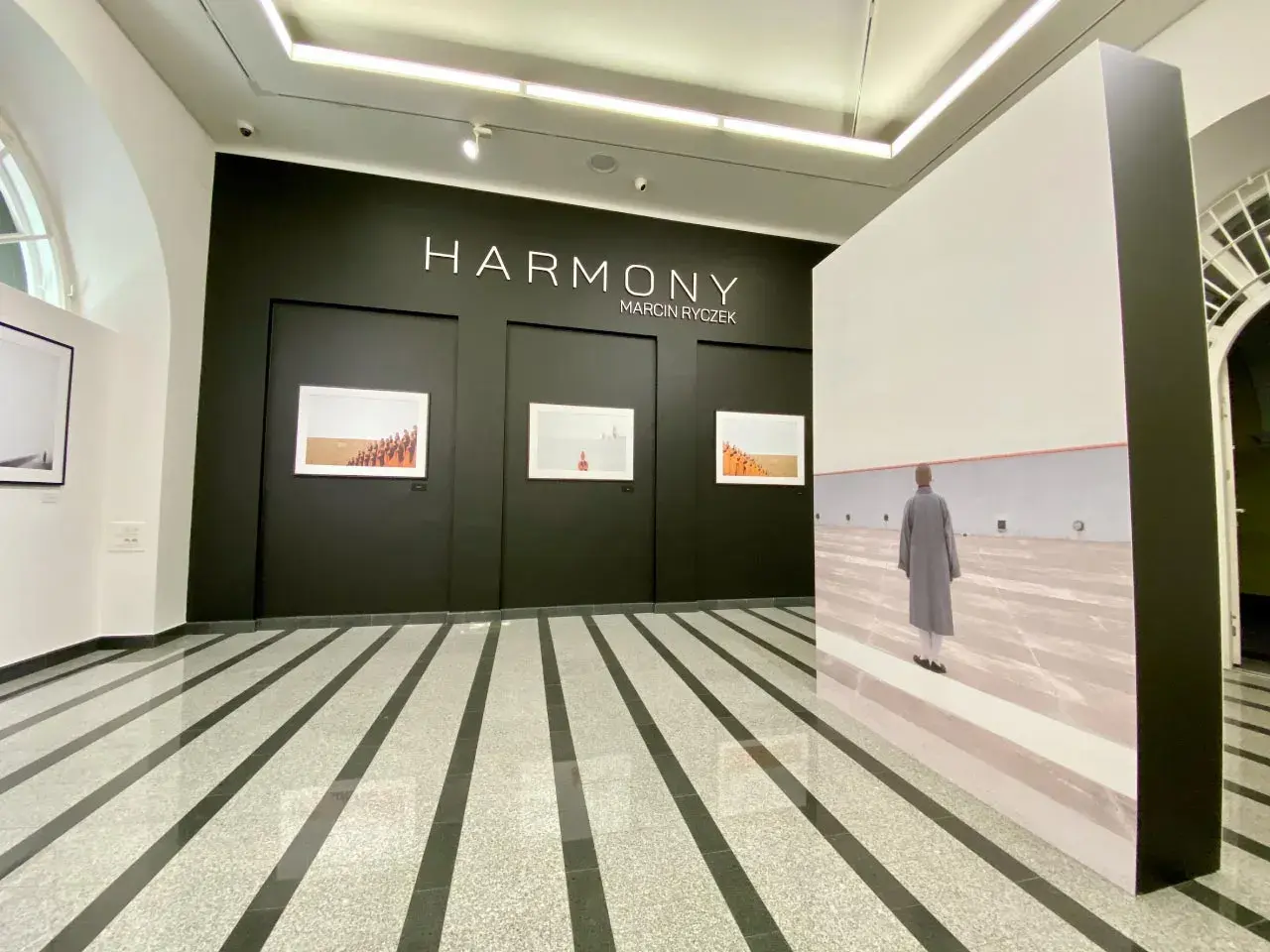 Wystawa "HARMONY" Marcina Ryczka. W galerii widzimy fotografie mnichów i dużą instalację z podobizną postaci w szarym płaszczu.