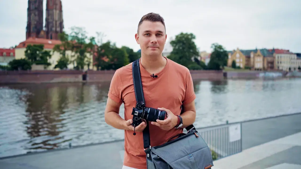 Mężczyzna z aparatem i torbą na ramieniu, gotowy do robienia zdjęć na dworze. W tle rzeka i zabytkowe budynki.