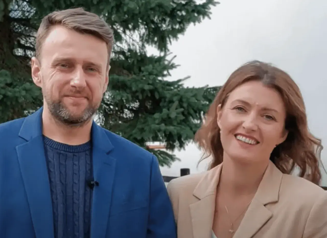 Uśmiechnięty adam kozioł w niebieskiej marynarce obok kobiety w beżowej.