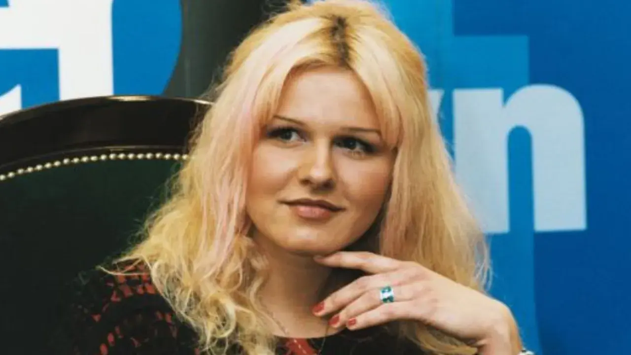Agnieszka Lorek z blond włosami i delikatnym uśmiechem, oparta o fotel, z ręką podtrzymującą podbródek.