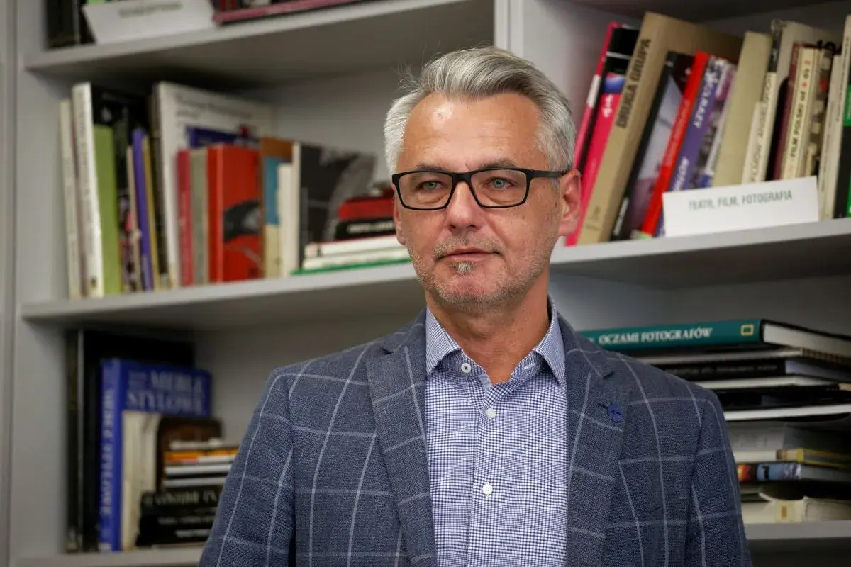 Andrzej Grygiel w okularach, w kratowaną marynarkę, na tle regałów z książkami o sztuce i fotografii.