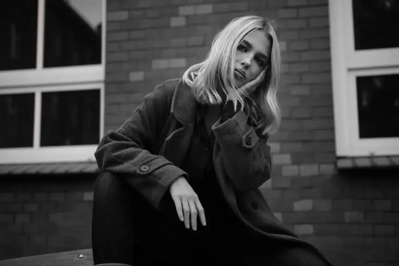 Blondynka w płaszczu opiera głowę na dłoni, zamyślona. Ten detal co to znaczy?