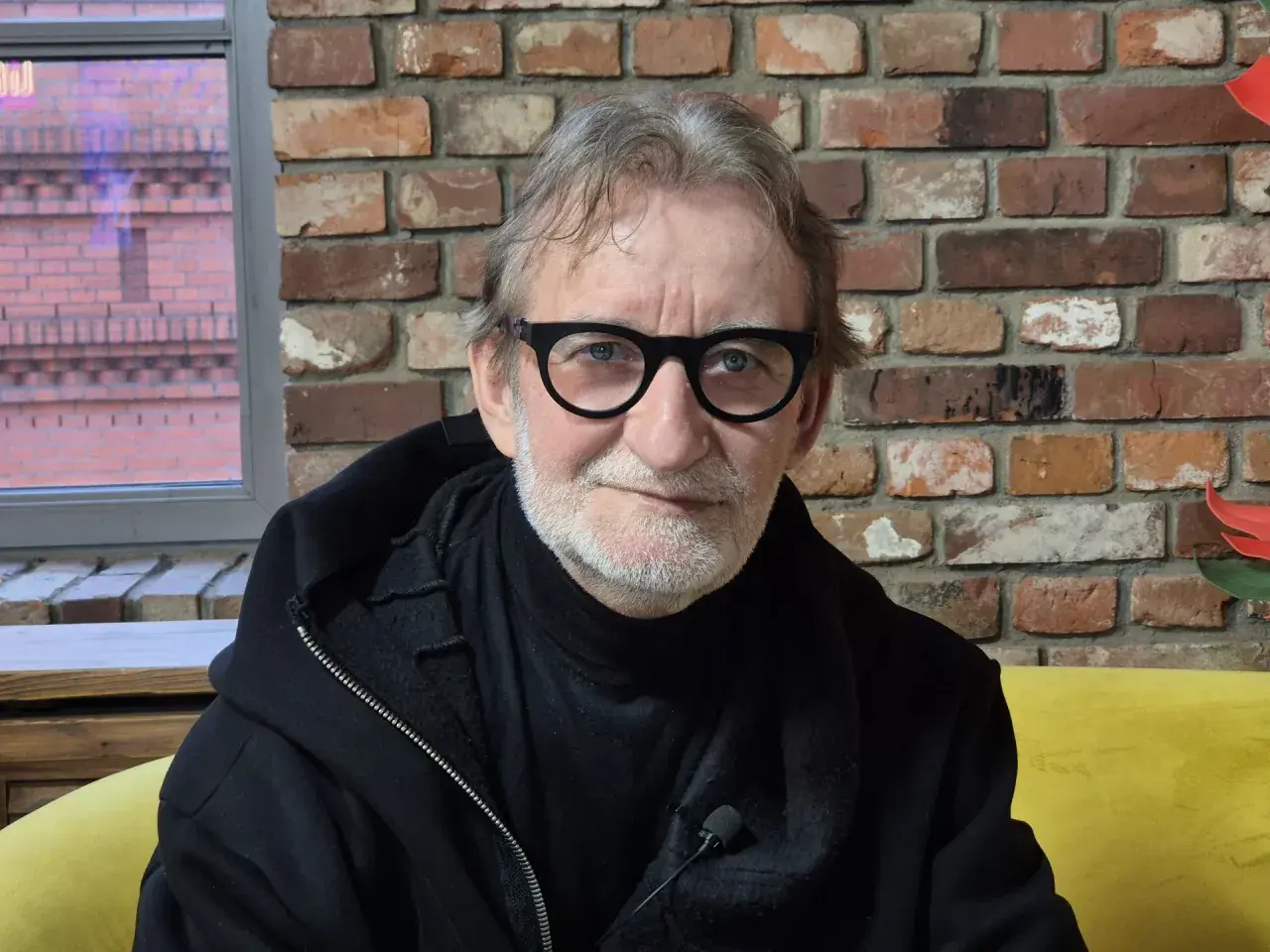 Maciej Mańkowski w okularach, z siwą brodą, w czarnej bluzie na tle ceglanej ściany.
