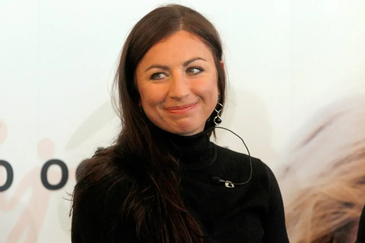 Uśmiechnięta Diana Kowalczyk w czarnym golfie, z charakterystycznymi kolczykami, patrzy w bok.