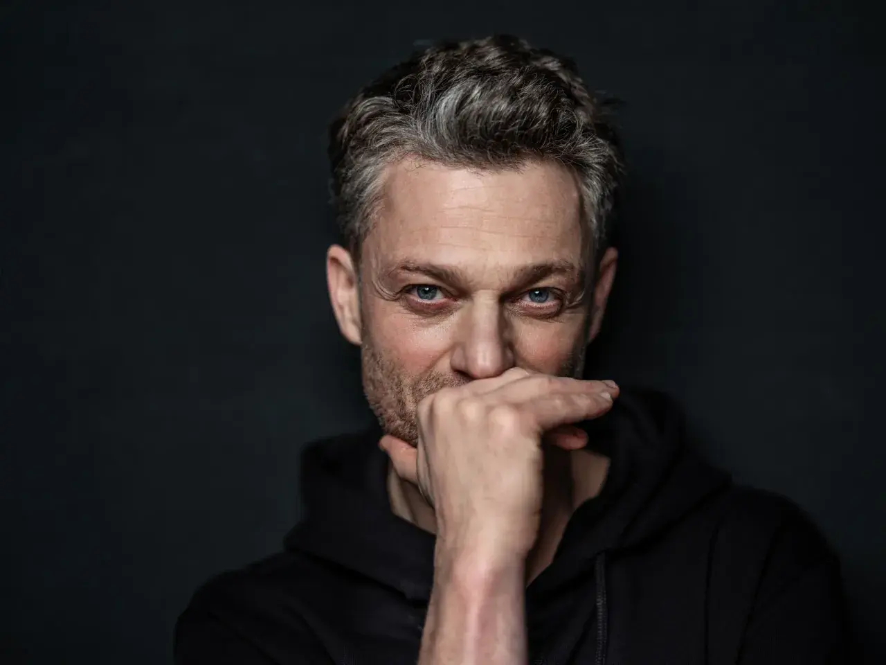 Szymon Szcześniak, fotograf, zamyślony, z dłonią na ustach, w czarnej bluzie z kapturem.