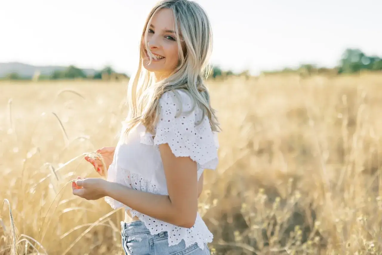 Uśmiechnięta blondynka podczas sesji na łące, trzymająca w dłoniach kłosy zbóż.