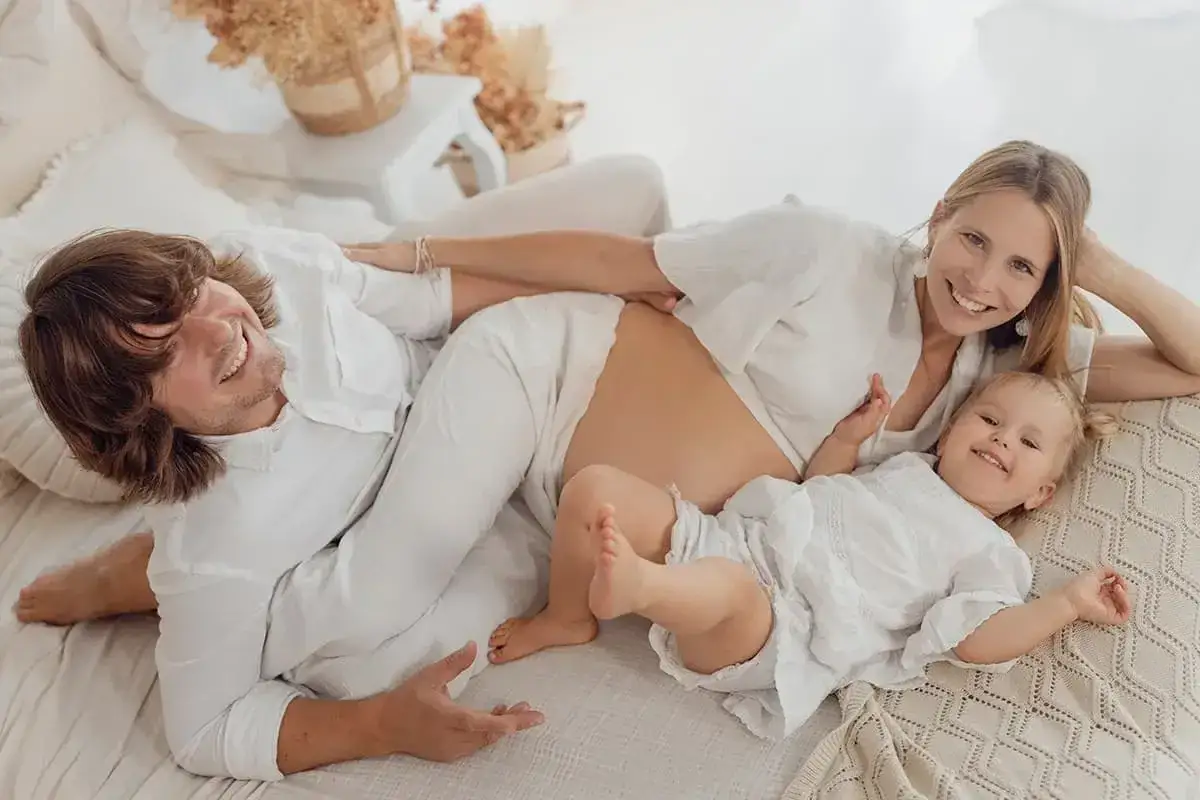 Szczęśliwa rodzina podczas sesji ciążowej. Uśmiechnięci rodzice i mała córeczka leżą na łóżku w białych ubraniach, celebrując oczekiwanie na nowego członka rodziny.