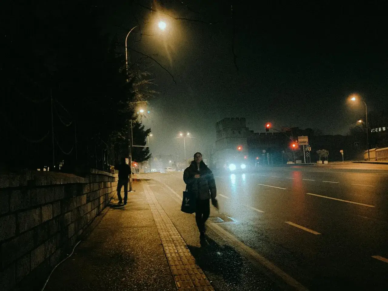 Nocne street foto. Kobieta idzie chodnikiem w deszczu, w tle światła miasta i zabytkowa budowla.