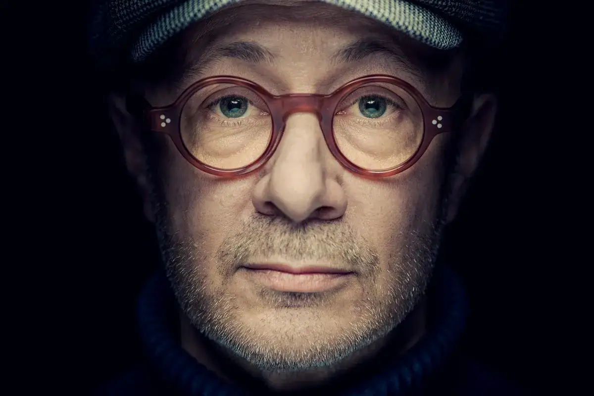 Portret Mikołaja Grynberga w okularach i czapce. Jego spojrzenie jest intensywne, a twarz oświetlona od przodu.