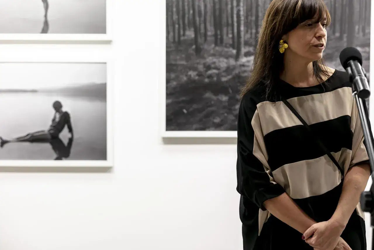 Joanna Chudy przemawia przy mikrofonie, w tle czarno-białe fotografie.