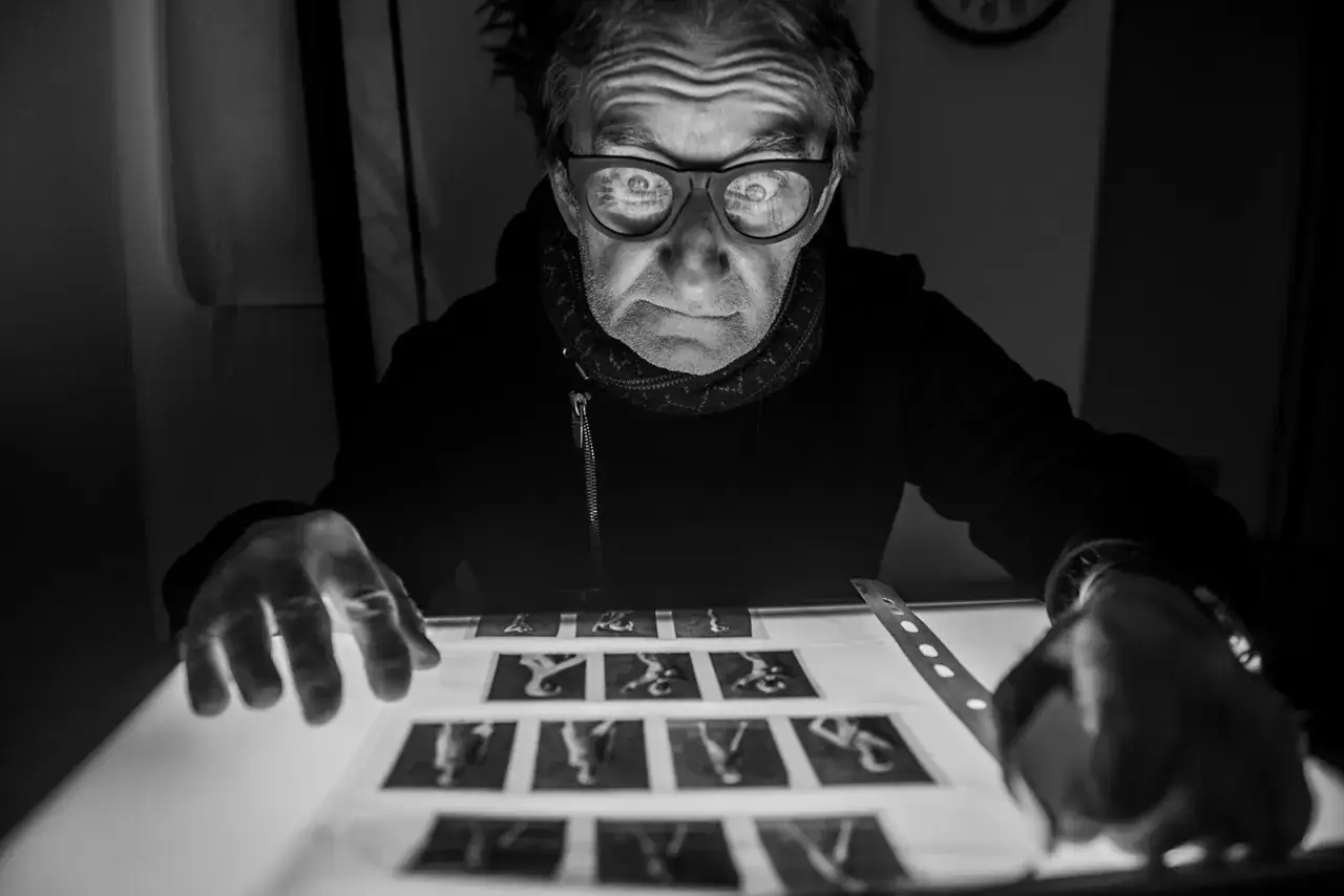 Maciej Mańkowski w okularach pochyla się nad negatywami naświetlanymi od spodu. Jego twarz jest pełna skupienia.