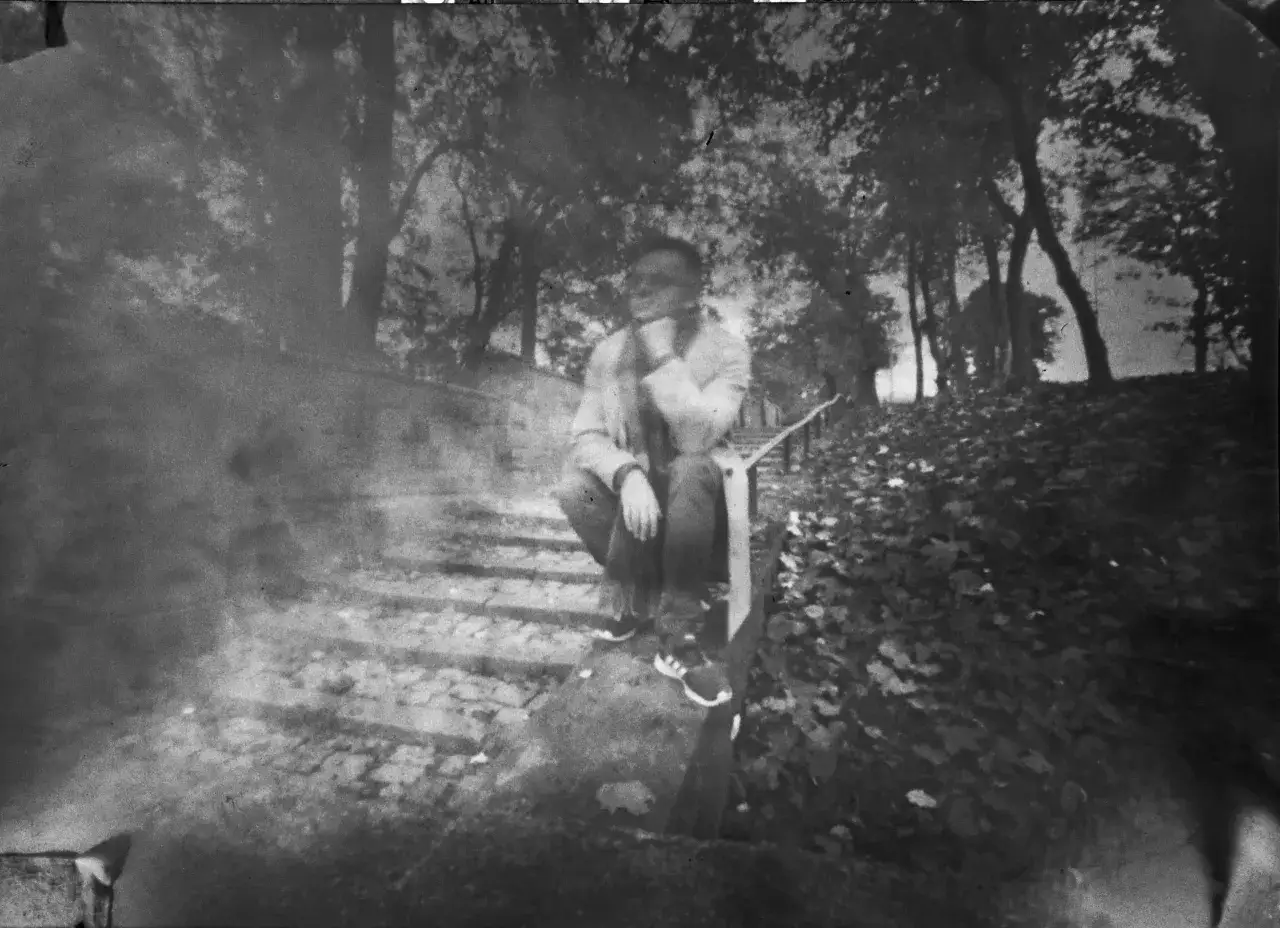 Osoba siedząca na schodach w parku, otoczona drzewami. Efekt starej fotografii otworkowej dodaje tajemniczości.