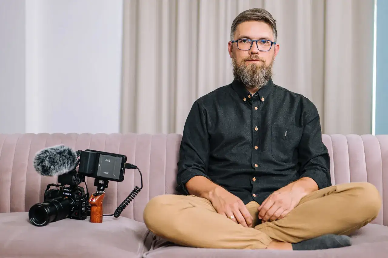 Krystian Bielatowicz, brodaty mężczyzna w okularach, siedzi na kanapie obok profesjonalnego sprzętu filmowego.