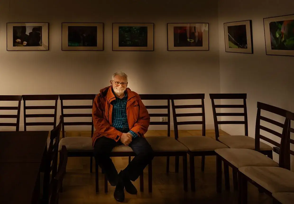 Uśmiechnięty Andrzej Zygmuntowicz siedzi na krześle w galerii, w tle wiszą obrazy.