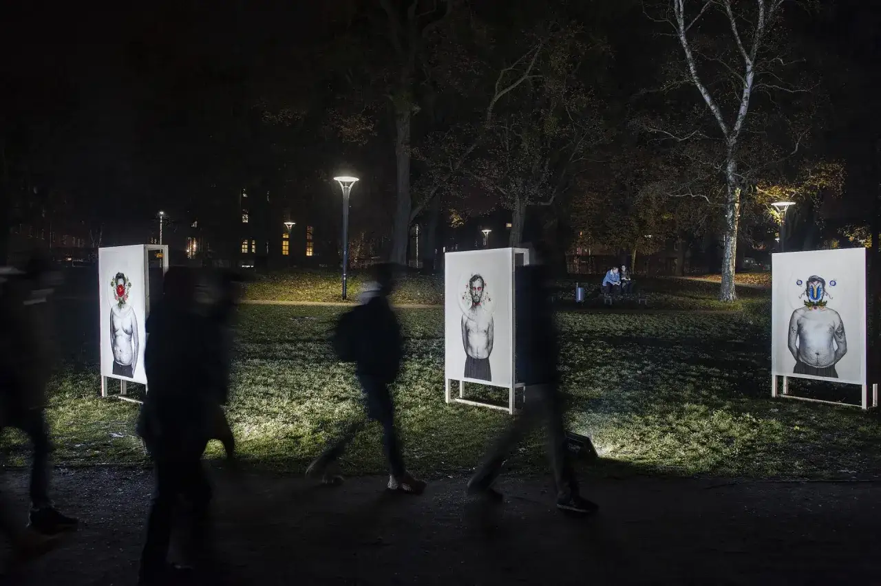 Nocna wystawa w parku. Portrety mężczyzn z artystycznymi nakryciami głowy, autorstwa Bartka Jakubowskiego, oświetlone reflektorami.