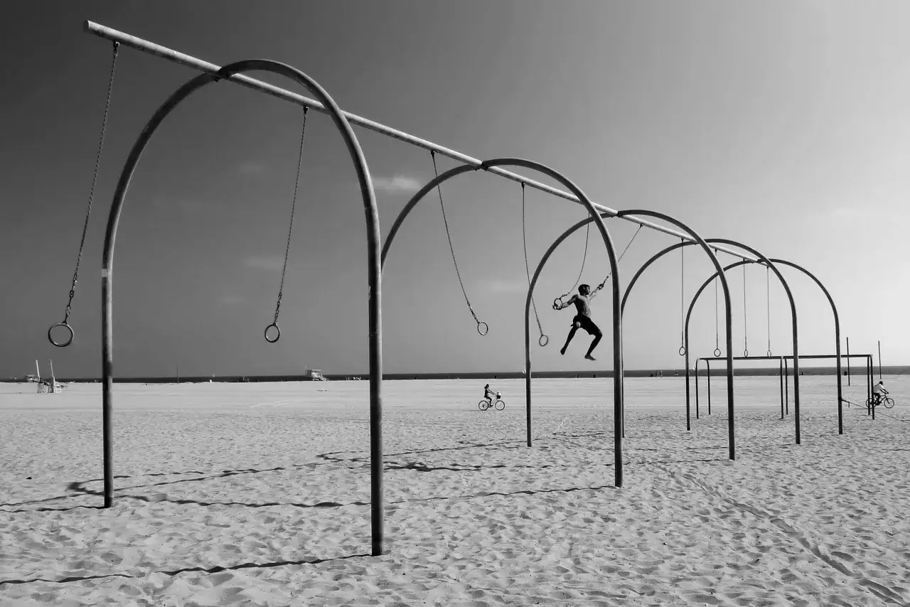 Kompozycja dynamiczna fotografia: człowiek wykonuje akrobacje na kółkach gimnastycznych na plaży, w tle rowerzysta.