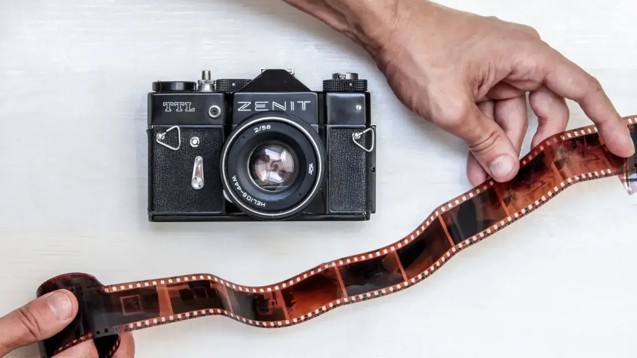 Dwie ręce trzymają kliszę filmową obok czarnego aparatu Zenit. To jakby przygotowanie do pracy w ciemni fotograficznej.