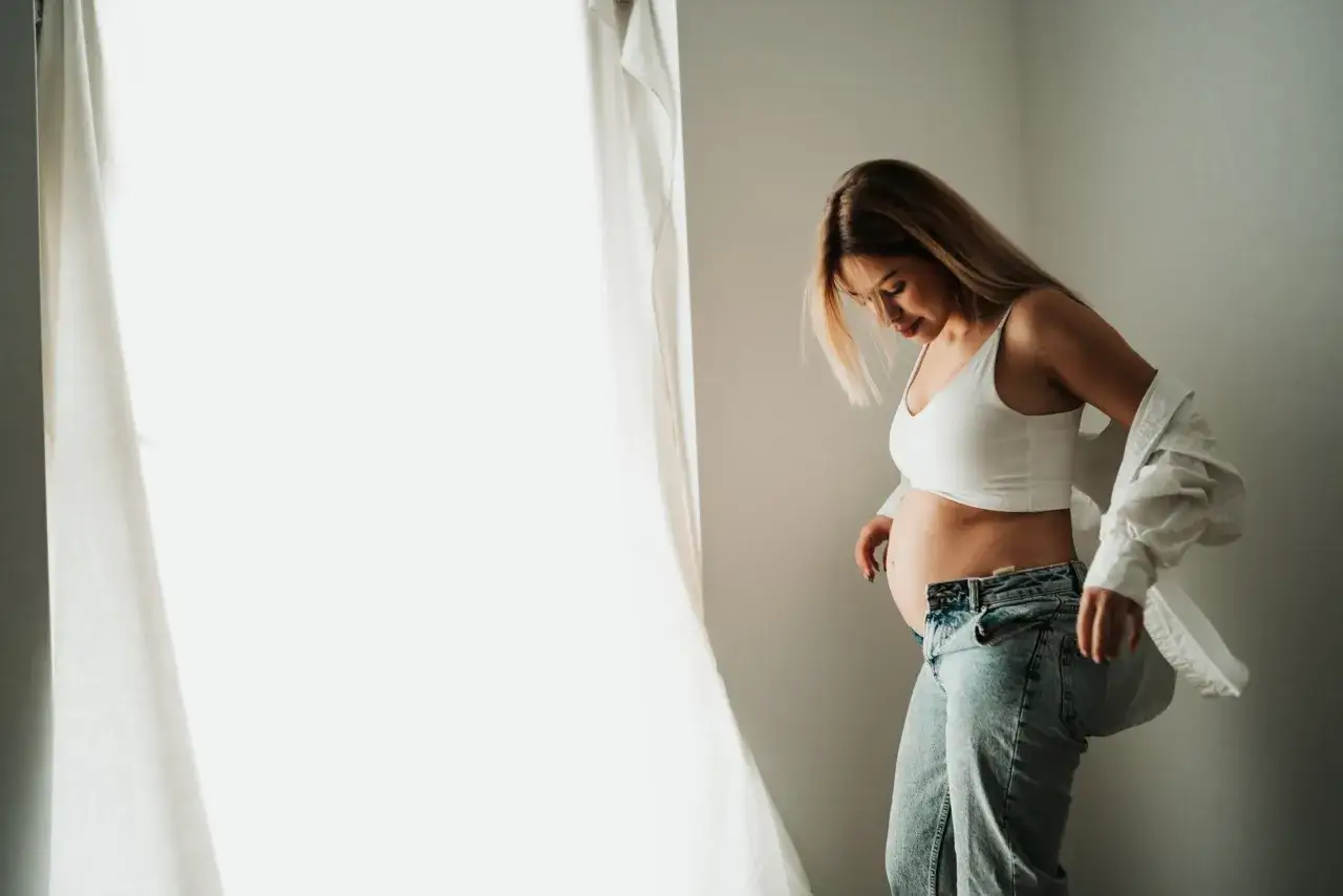 Kobieta w białym topie i jeansach, z dłonią na brzuchu. Naturalne światło podkreśla jej ciążowy brzuszek. Świetne pomysły na sesję ciążową w domu.