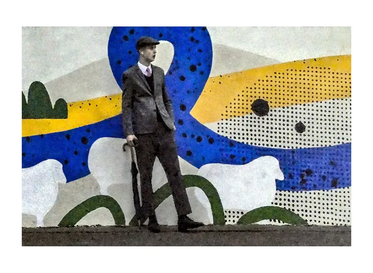 Tomek Sikora w eleganckim garniturze i kaszkiecie, z parasolką, na tle abstrakcyjnego malowidła z owcami i geometrycznymi wzorami.