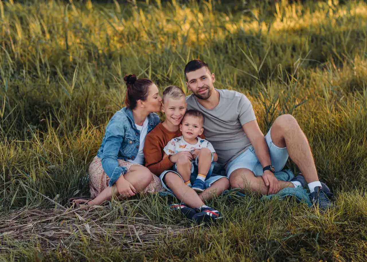 Rodzina podczas sesji lifestyle na łące. Mama całuje syna, który siedzi na kolanach taty. Obok starszy brat.
