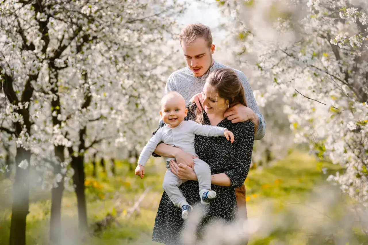 Uśmiechnięta rodzina w kwitnącym sadzie. Mama trzyma dziecko, a tata obejmuje ich. To była naprawdę śmieszna sesja rodzinna.