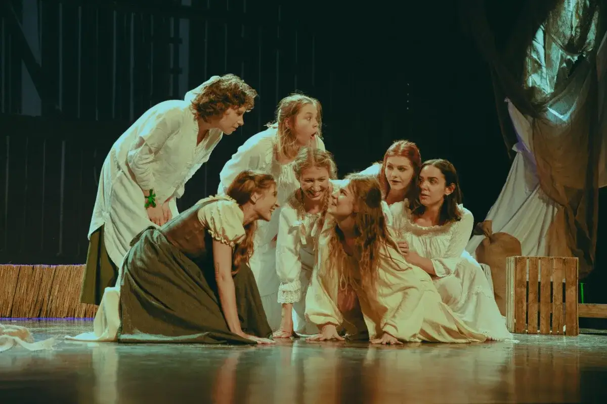 Grupa młodych kobiet w strojach z epoki na scenie. Ta fotografia teatralna uchwyciła emocje i dynamikę sceny.