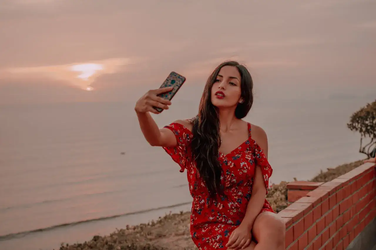 Kobieta w czerwonej sukience z kwiatami robi sobie selfie na tle zachodzącego słońca nad morzem. Idealny pomysł na selfie.