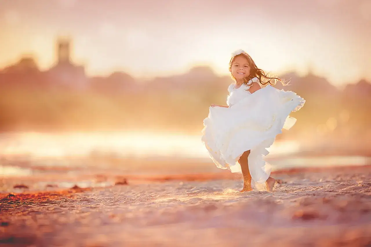 Dziewczynka w białej sukience tańczy na plaży podczas złotej godziny w fotografii. W tle widać zarys budowli.