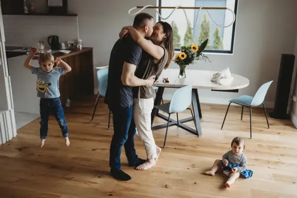 Rodzice w uścisku, starszy syn skacze, młodsza córka siedzi na podłodze. Urocza sesja lifestyle w domu.