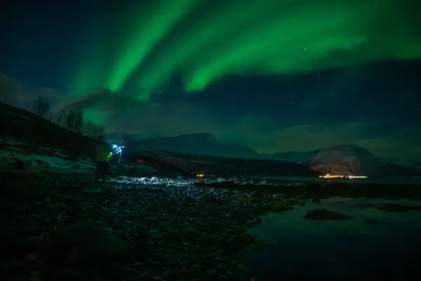 Nocne niebo rozświetla zorza polarna. Na pierwszym planie widać fotografa z lampą, który robi zdjecie z fleszem.