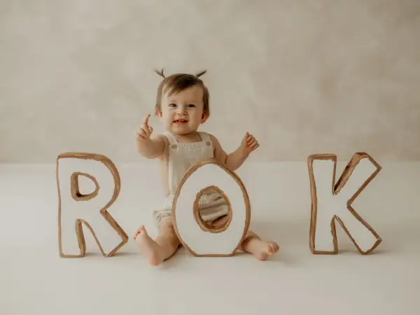 Uśmiechnięte dziecko z dwoma kucykami świętuje pierwszy roczek podczas sesji retro. Litery R, O, K tworzą napis "ROK".