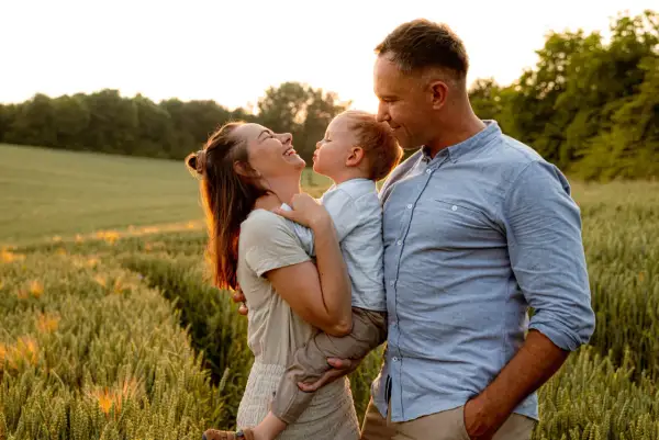 Szczęśliwa rodzina podczas sesji na łące. Mama śmieje się, trzymając dziecko, a tata patrzy na nich z czułością.