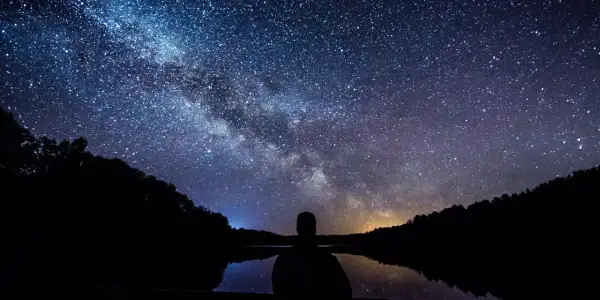 Sylwetka człowieka patrzy na Drogę Mleczną nad jeziorem. Idealny kadr, by pokazać, jak fotografować gwiazdy.