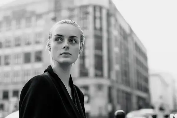 Monochromatyczna fotografia. Młoda kobieta o blond włosach patrzy w bok, na tle miejskiej architektury.