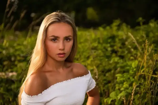Młoda kobieta z długimi blond włosami, ubrana w białą, odkrywającą ramiona bluzkę, patrzy prosto w obiektyw na tle zielonej, rozmytej roślinności.