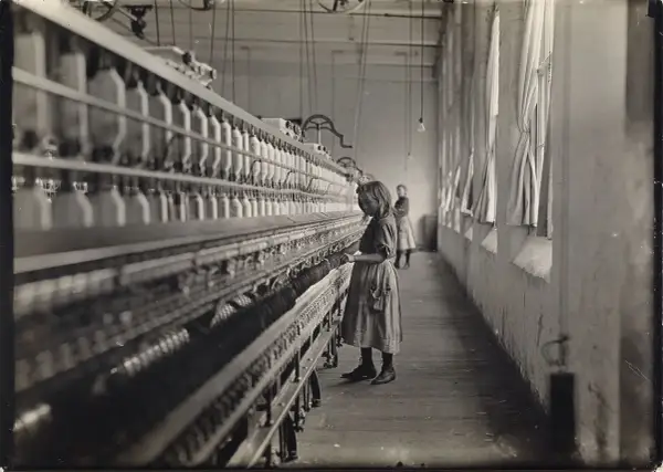 Dziewczynka pracuje przy maszynie w fabryce, jak na zdjęciach Lewis Hine. W tle widać inną pracownicę.