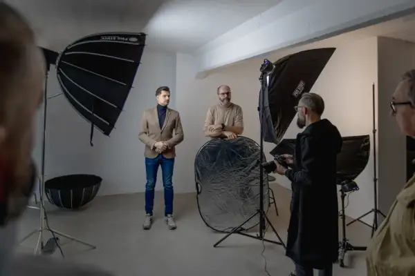 W studiu fotograficznym trwa sesja. Widać ludzi, sprzęt i modyfikatory światła w fotografii, w tym parasole i blendę.