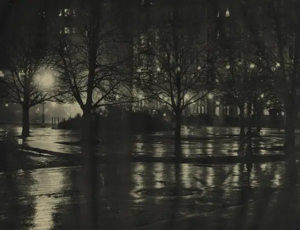 Nocny park po deszczu, drzewa bez liści, mokre ścieżki odbijające światła budynków. Alfred Stieglitz uchwycił atmosferę miasta.