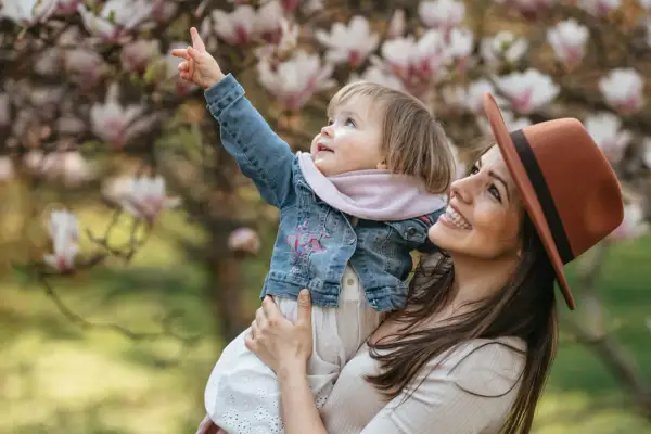 Uśmiechnięta mama w kapeluszu trzyma dziecko podczas sesji wiosennej dzieci. Maluch wskazuje palcem na kwitnącą magnolię.