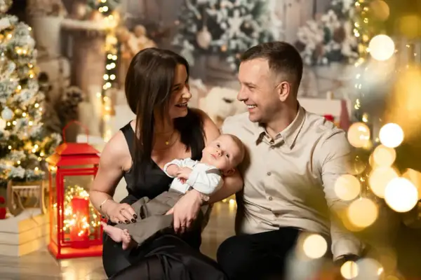 Rodzina podczas sesji bożonarodzeniowych. Mama i tata patrzą na siebie z uśmiechem, trzymając niemowlę. W tle choinki i lampki.