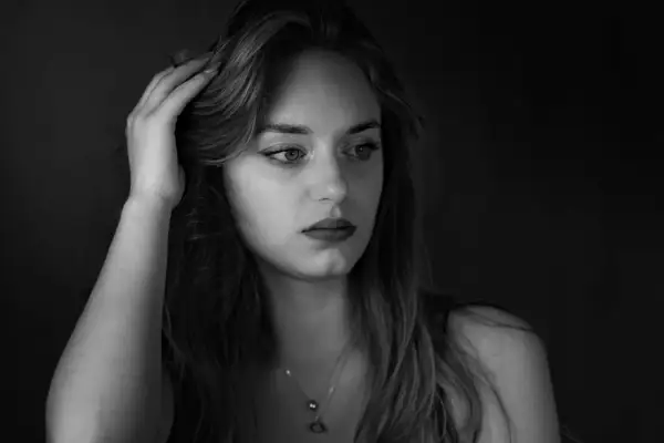 Portret młodej kobiety, fotografia czarno biała. Jej dłoń spoczywa na włosach, wzrok skierowany w bok, w zamyśleniu.