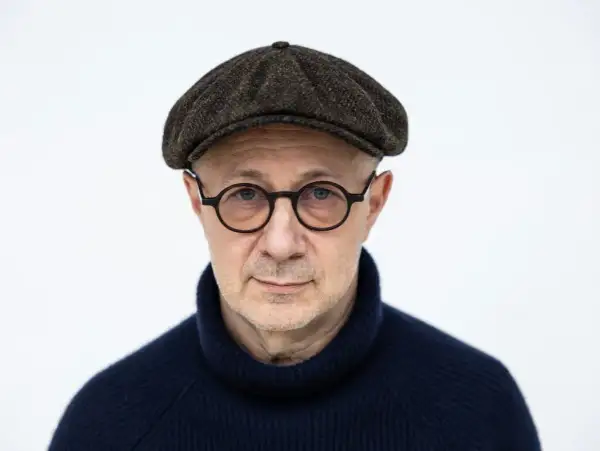 Mikołaj Grynberg w okularach i kaszkiecie, zamyślony, w granatowym golfie.