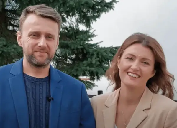 Uśmiechnięty adam kozioł w niebieskiej marynarce obok kobiety w beżowej.