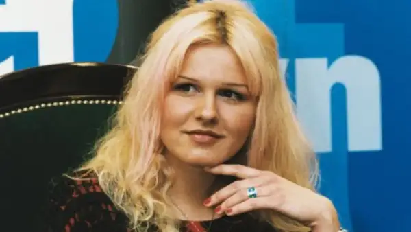 Agnieszka Lorek z blond włosami i delikatnym uśmiechem, oparta o fotel, z ręką podtrzymującą podbródek.
