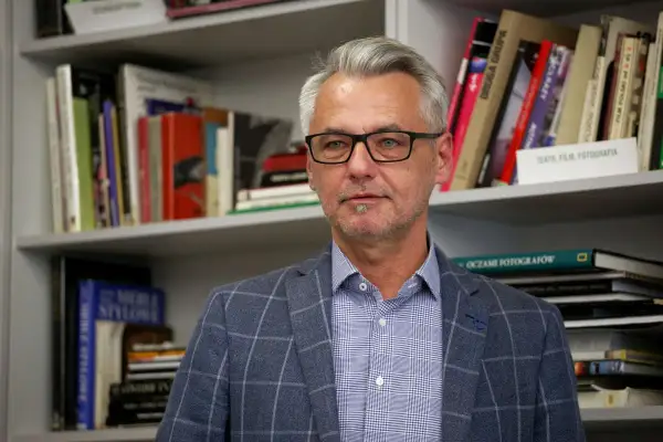 Andrzej Grygiel w okularach, w kratowaną marynarkę, na tle regałów z książkami o sztuce i fotografii.