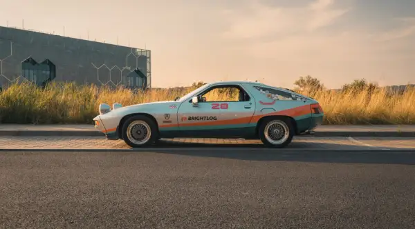 Klasyczna fotografia samochodowa: Porsche 928 w kolorach retro, z numerem 28 i logiem Brightlog, na tle nowoczesnego budynku i złotej trawy.