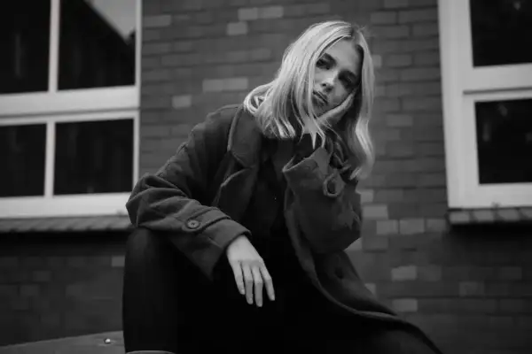 Blondynka w płaszczu opiera głowę na dłoni, zamyślona. Ten detal co to znaczy?